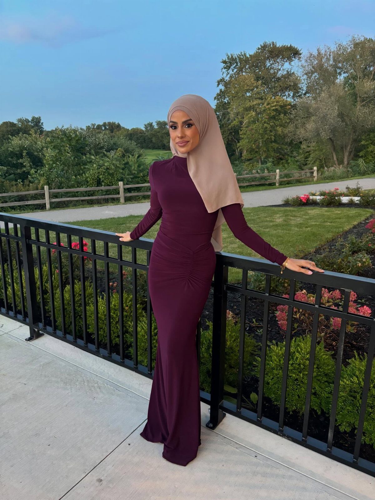Woman in a long-sleeved plum crepe dress and tan hijab posing by a black railing.
