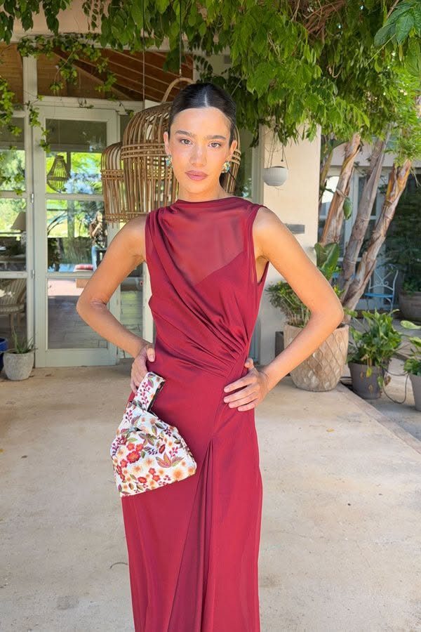 Woman in a sleeveless ruby red draped dress posing outdoors with a floral clutch.