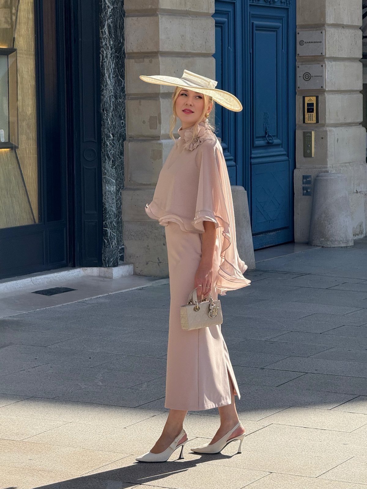 A woman in a pale pink outfit and wide-brimmed hat posing on a city street.
