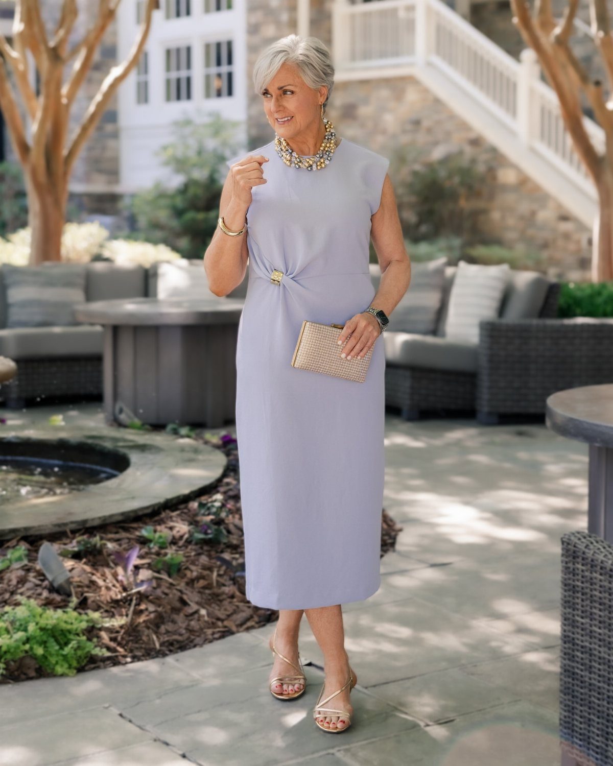 A mature woman posing in a sleeveless lavender midi dress and gold accessories outdoors.