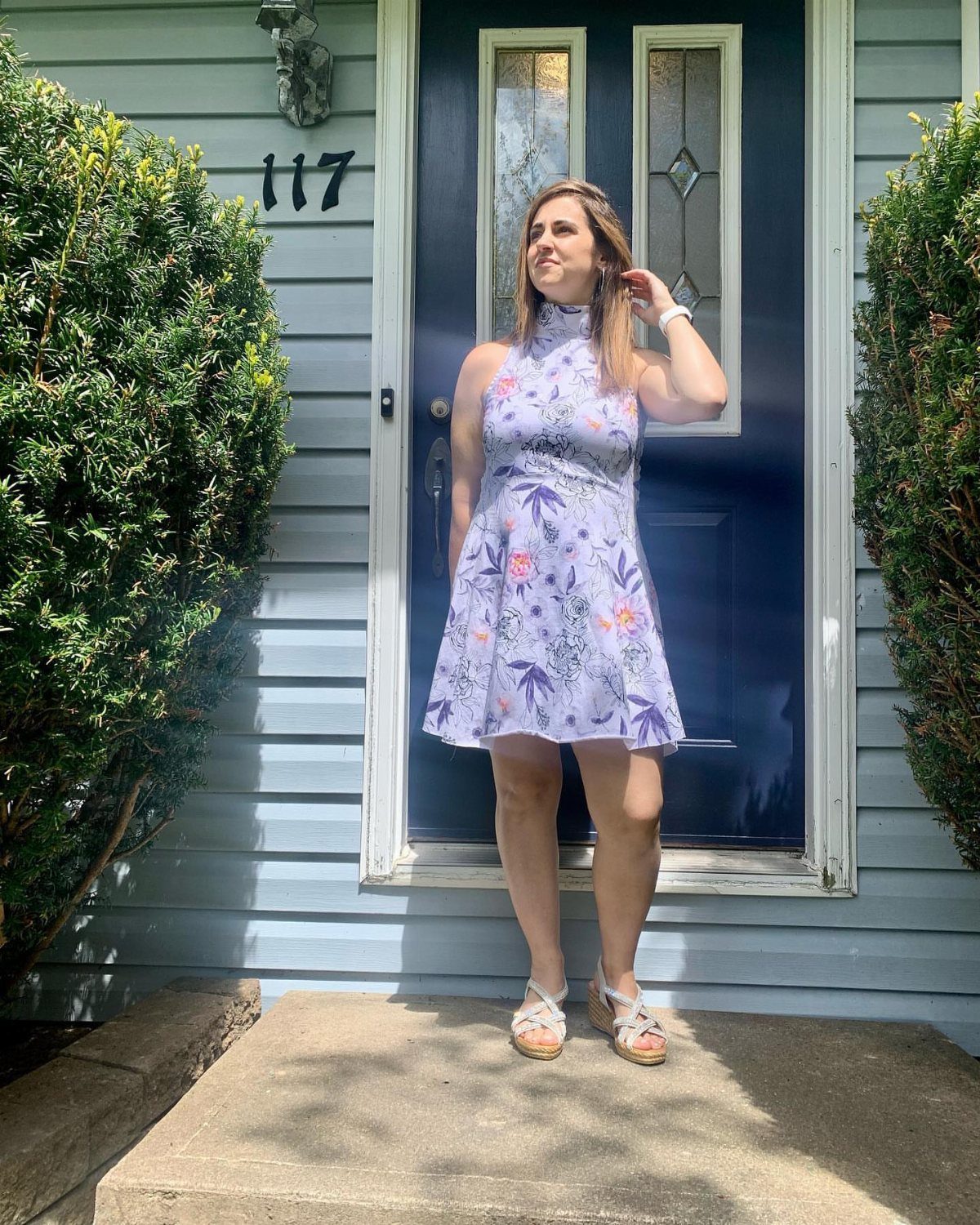 A woman in a purple floral halter dress and white sandals standing by a blue door.