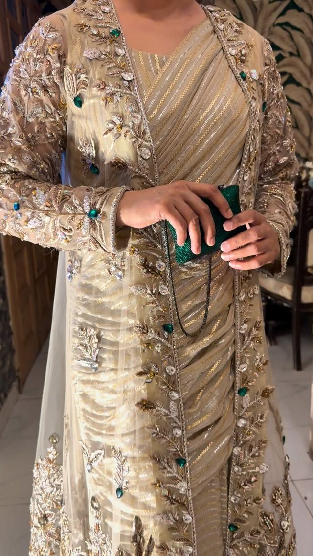 Person wearing an embellished gold gown and a sheer, floral embroidered jacket holding a green clutch.