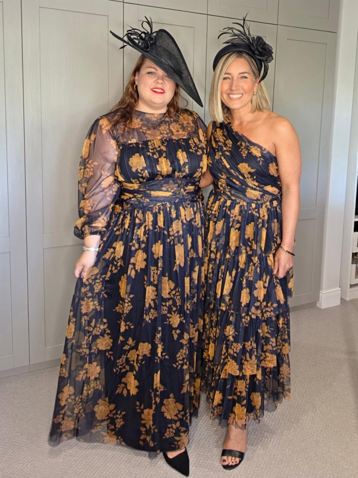 Two women in matching navy and gold floral dresses and black fascinator hats.