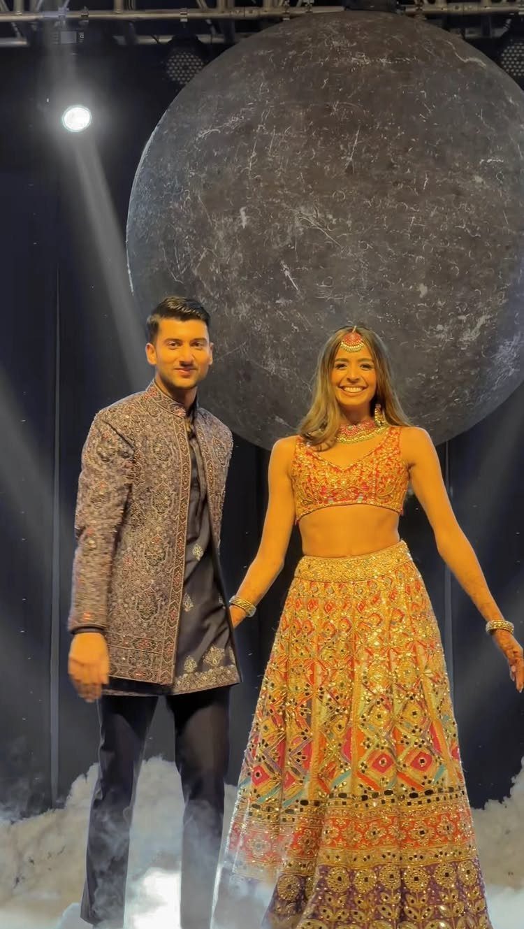 A man and a woman in colorful traditional Indian attire posing against a large moon backdrop.