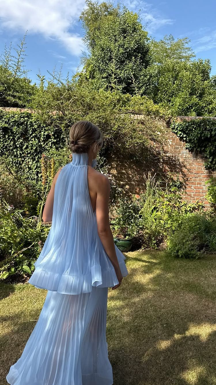 Woman in a light blue pleated halter-neck gown standing in a lush green garden.