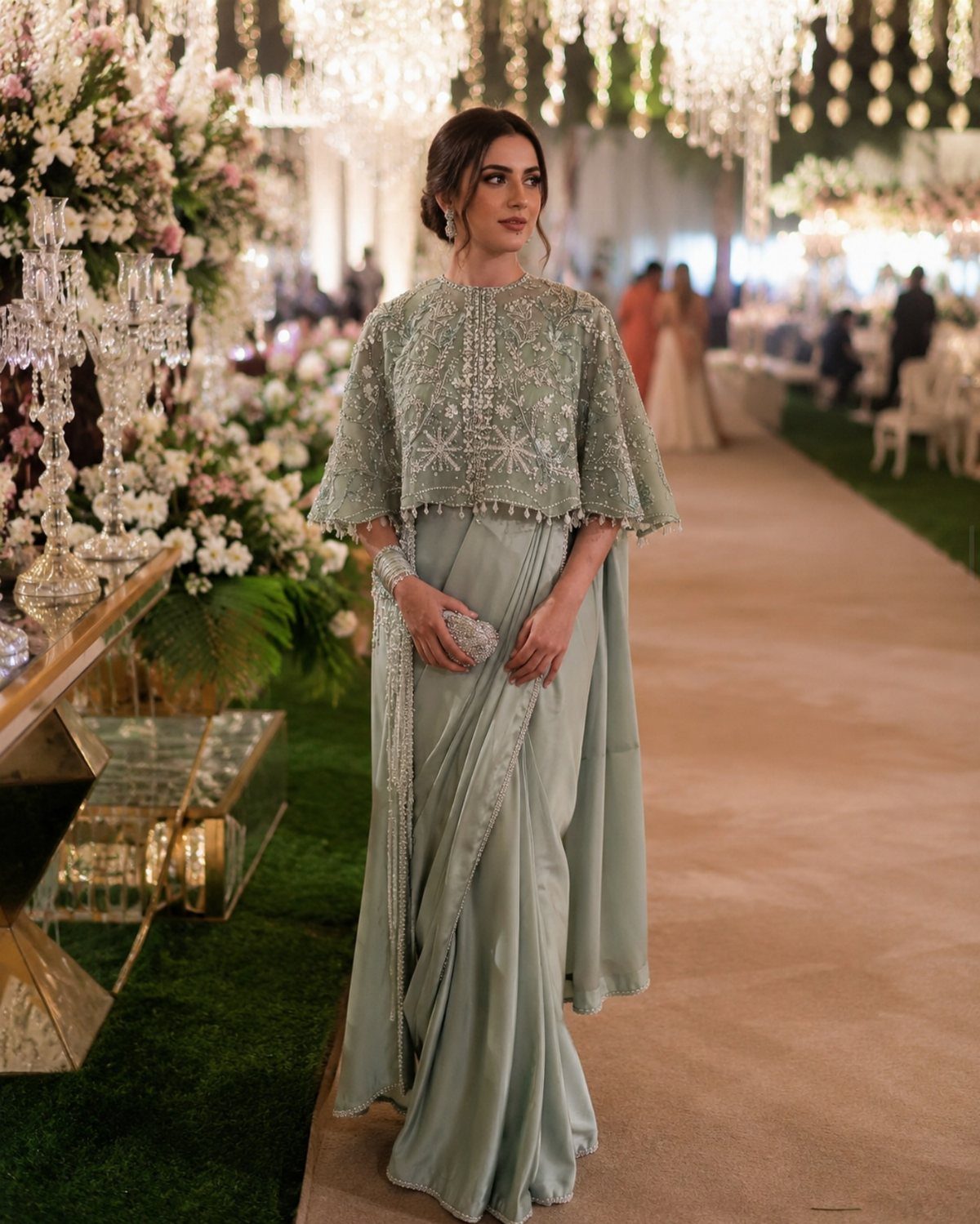 Woman in an elegant sage green embellished saree and lace cape standing in a decorated garden.