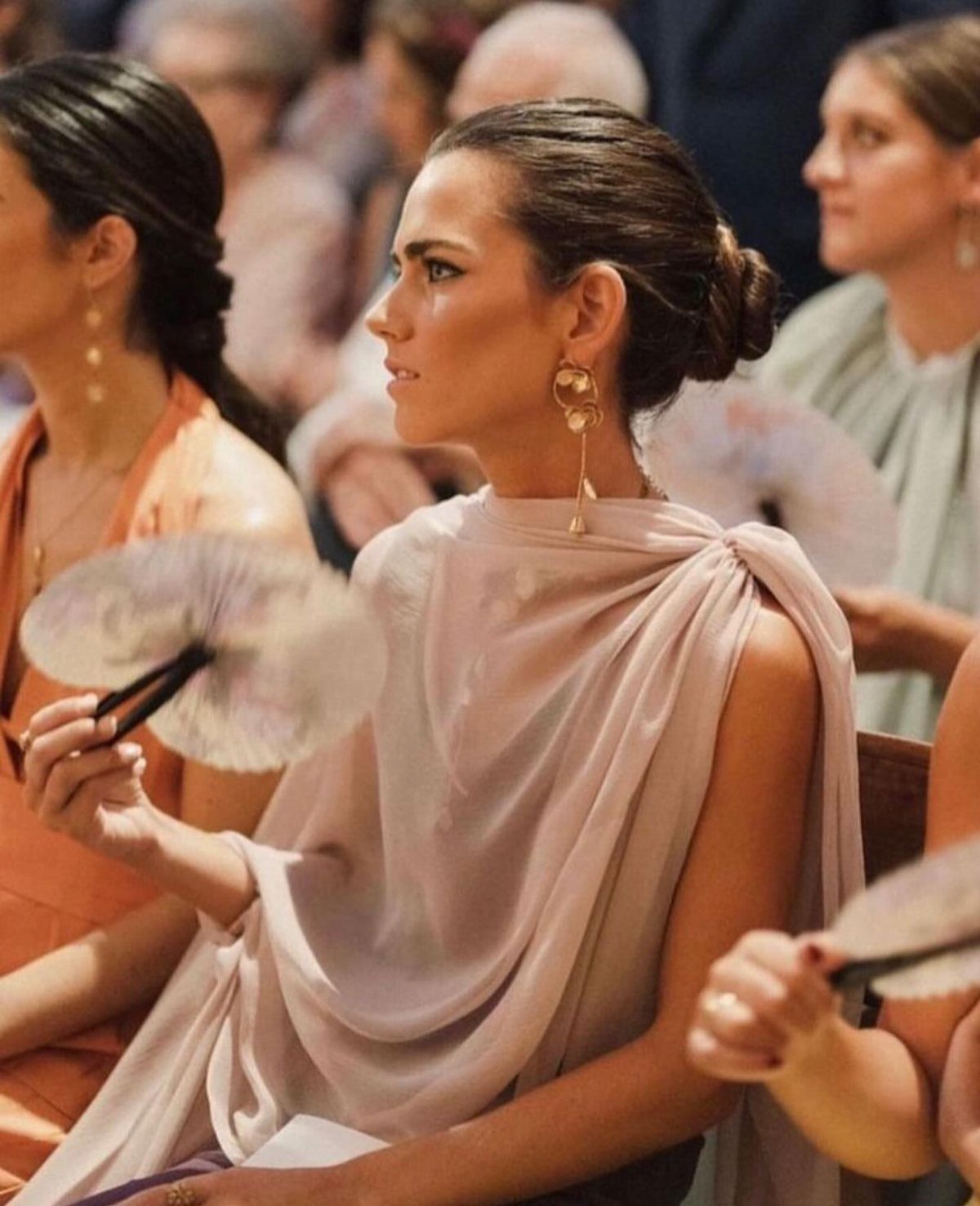Woman in a lavender draped dress wearing large gold statement earrings while holding a white fan.