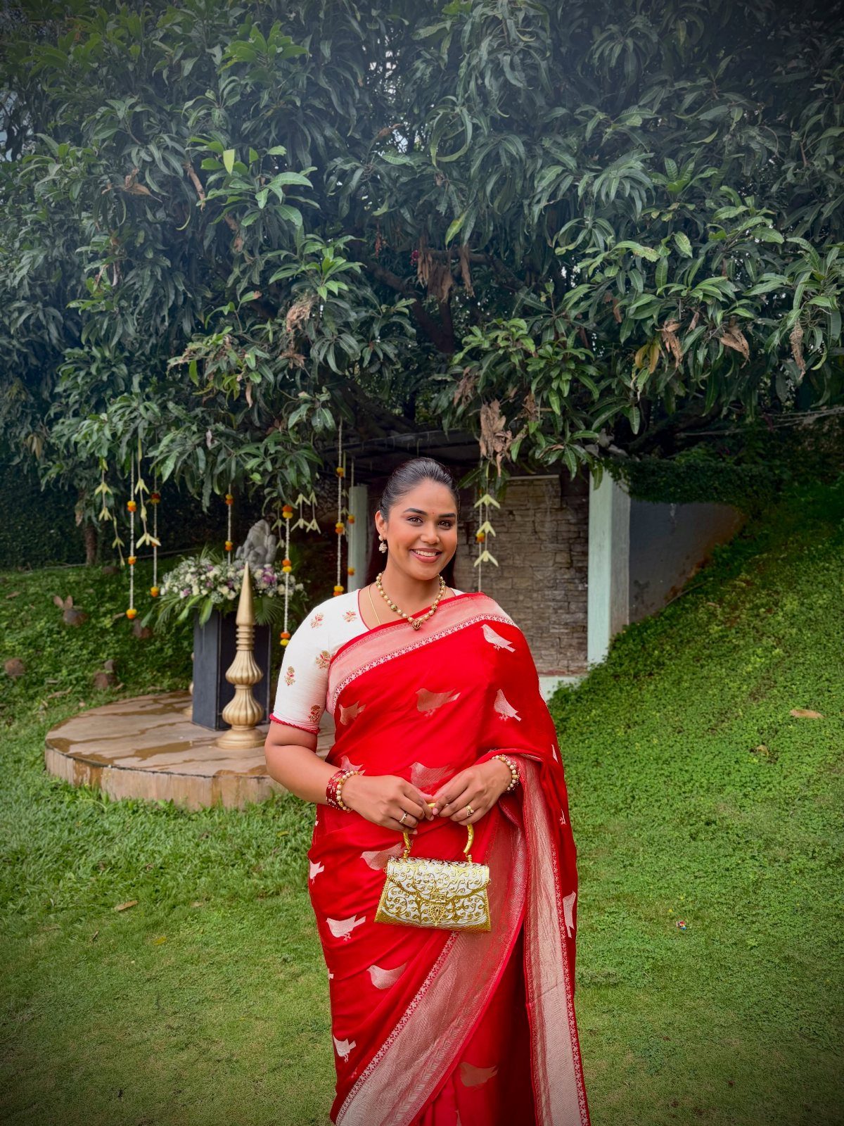 A woman in a red silk saree posing in a green garden setting.
