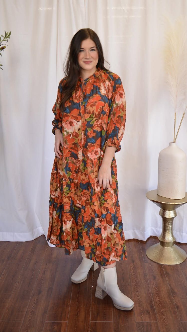 A woman wearing a long floral print dress and white ankle boots standing against white curtains.
