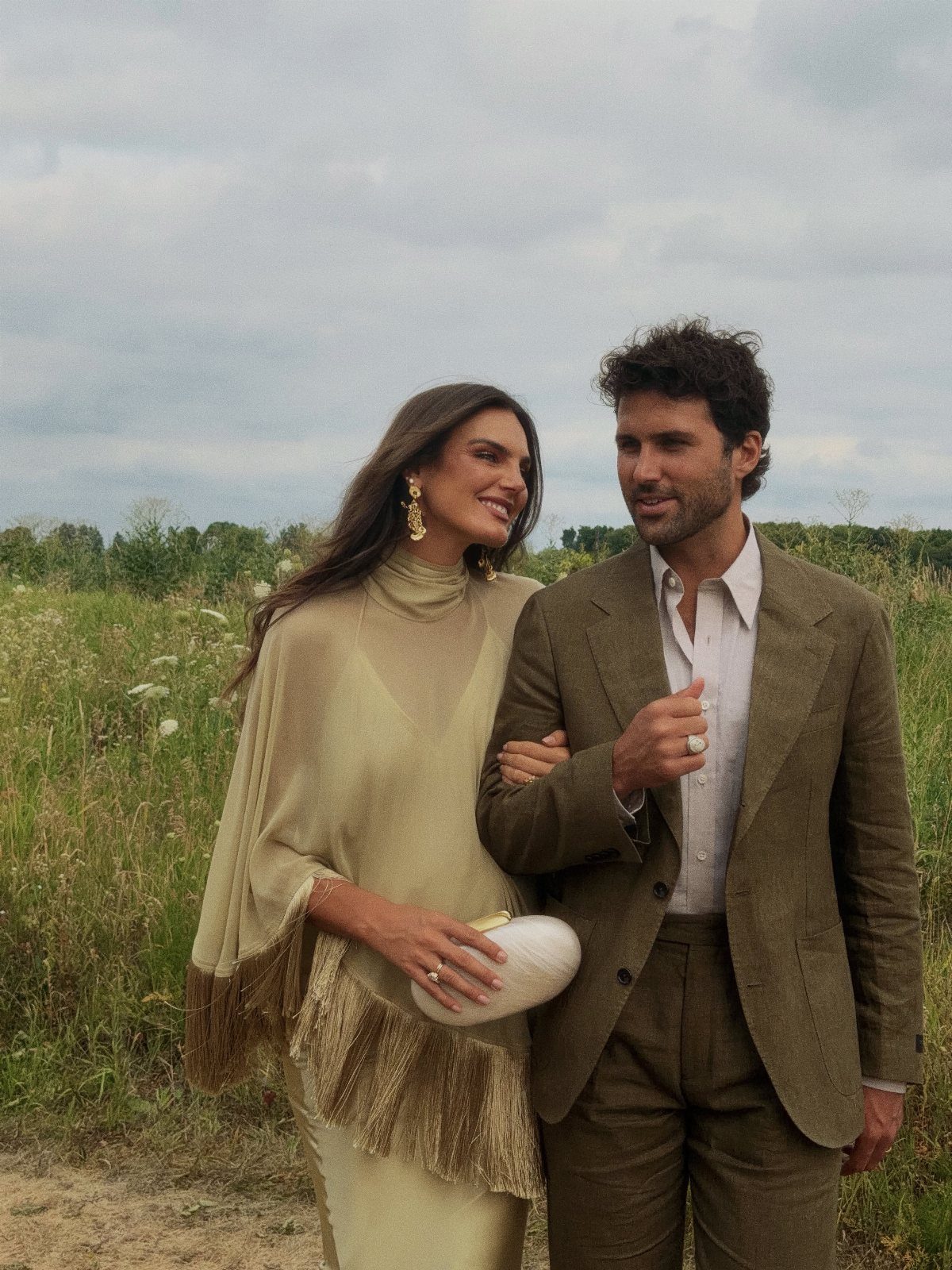 A man in a brown suit and a woman in a fringed olive green dress standing in a field.