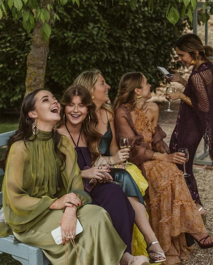 Five women in elegant dresses sitting on a bench outdoors, laughing and talking together.