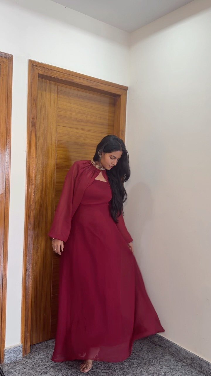 Woman in a long burgundy gown with flowing sleeves standing near a wooden door.