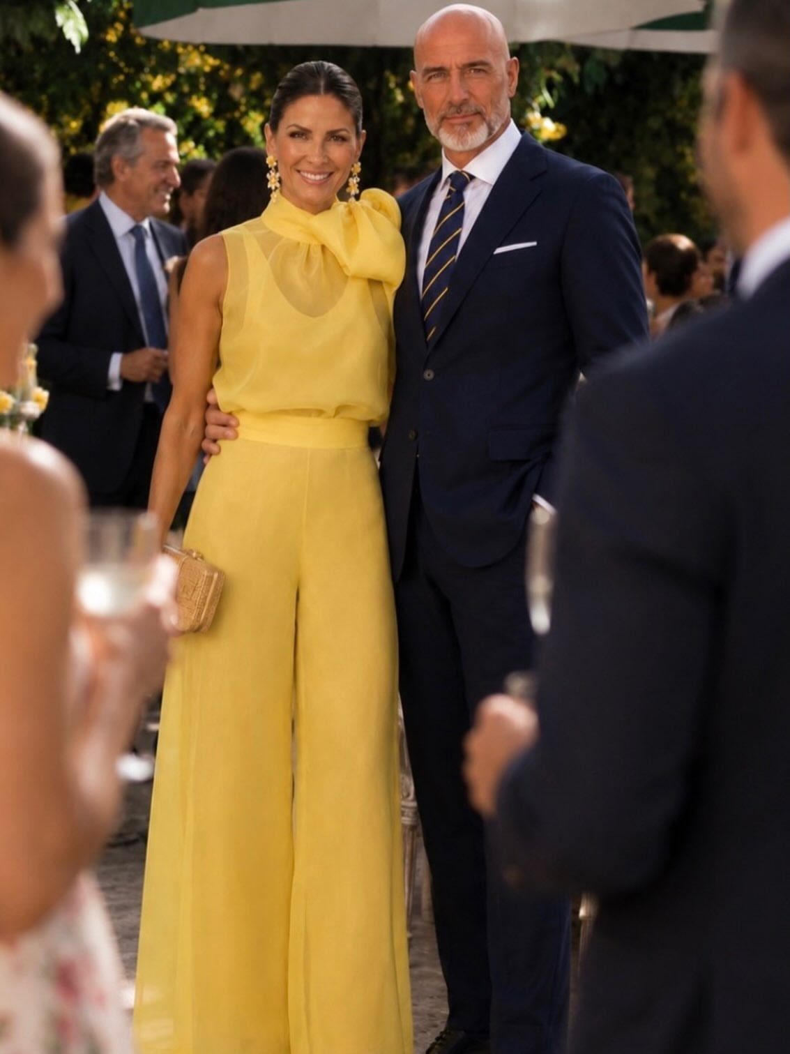 A woman wearing a bright yellow sleeveless jumpsuit with a bow detail stands smiling outdoors.
