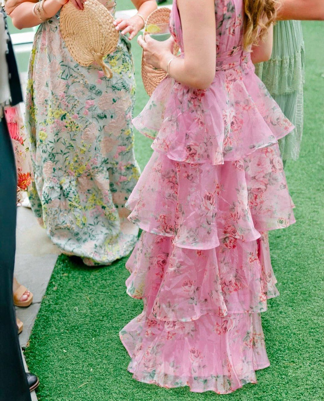 Women in long floral dresses standing on green grass holding straw fans.