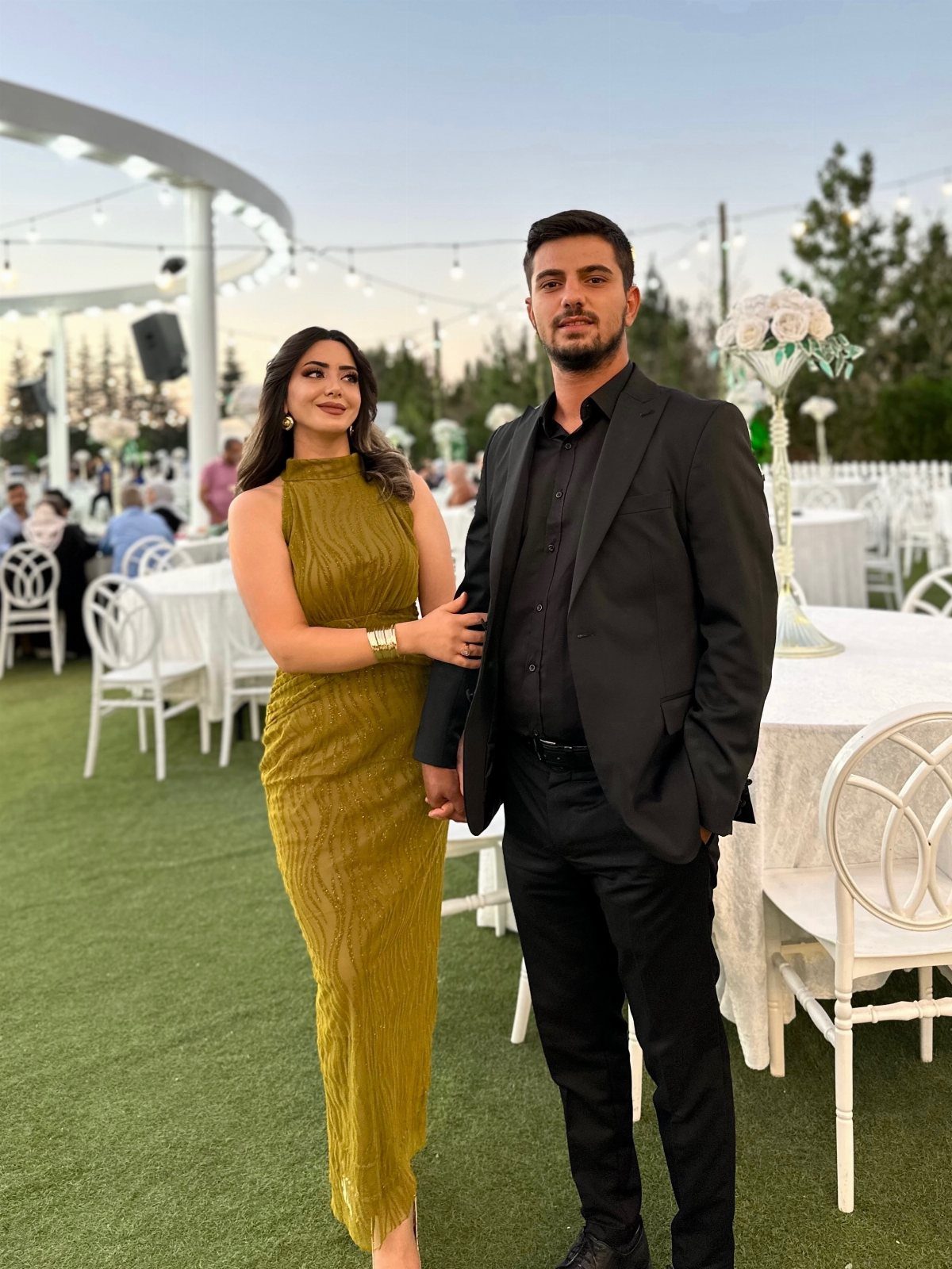 A man in a black suit and a woman in an olive green dress posing outdoors.