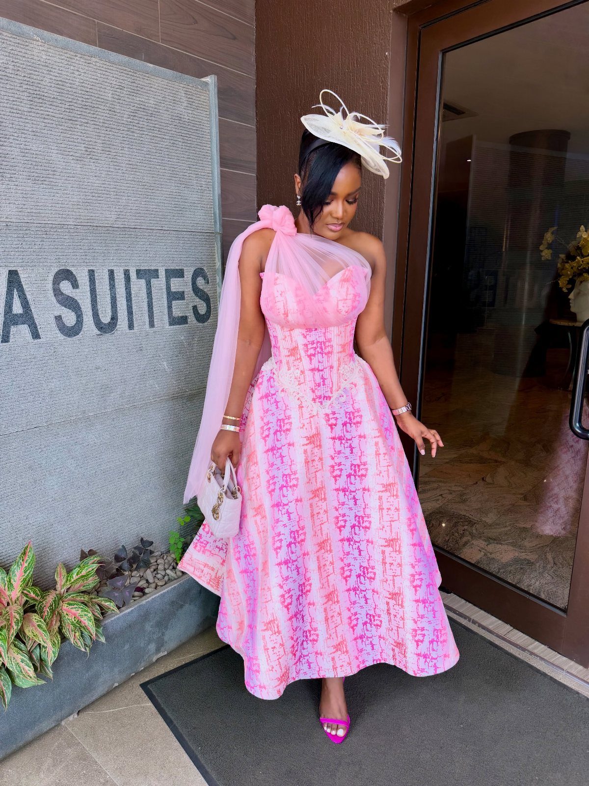 Woman in a pink patterned dress posing with a white fascinator and small handbag.