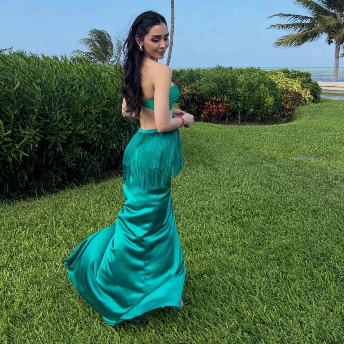 Woman in a teal satin mermaid gown posing on a tropical lawn near the ocean.