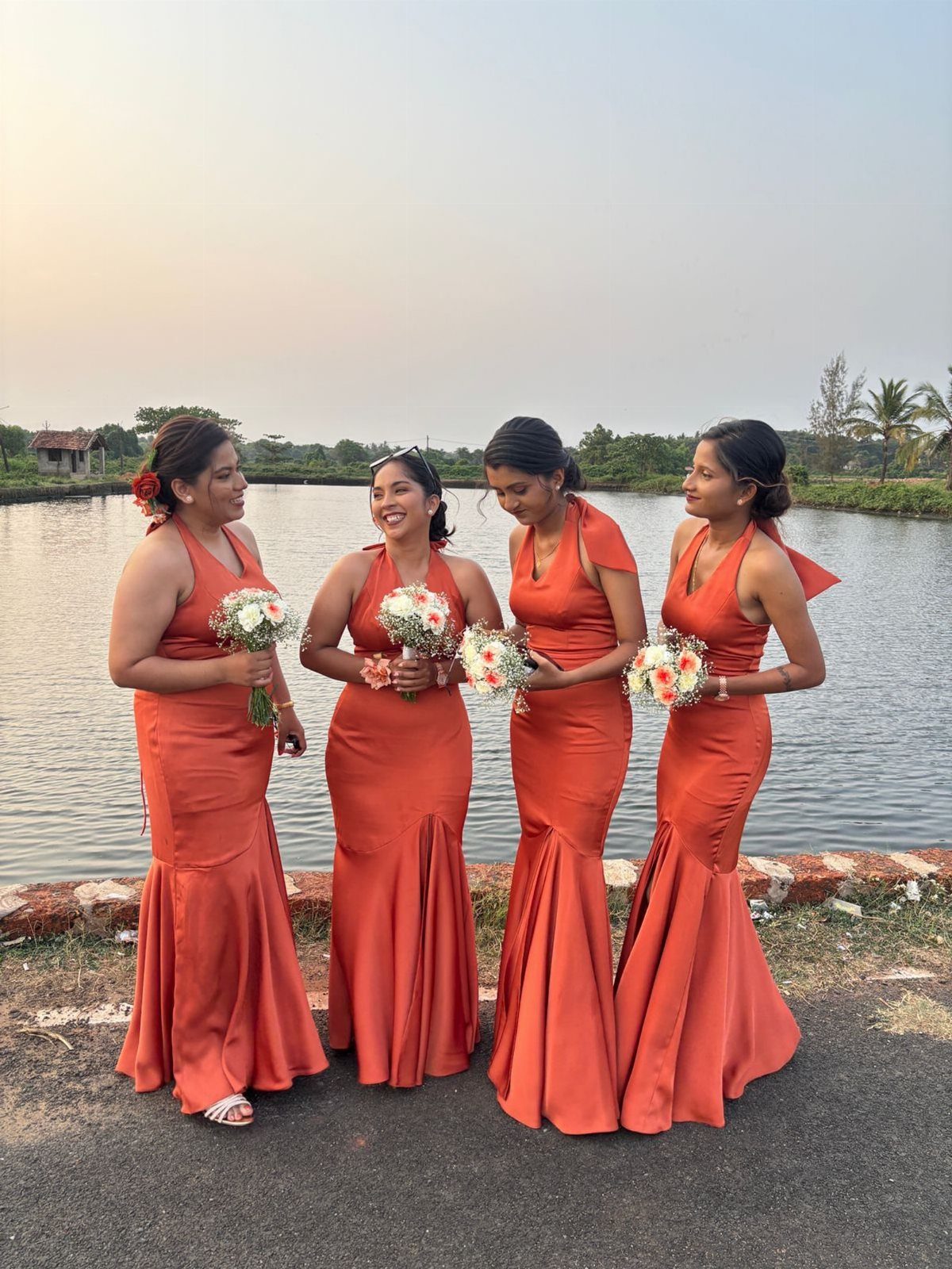 Four bridesmaids in burnt orange satin dresses holding small bouquets by a lake at sunset.