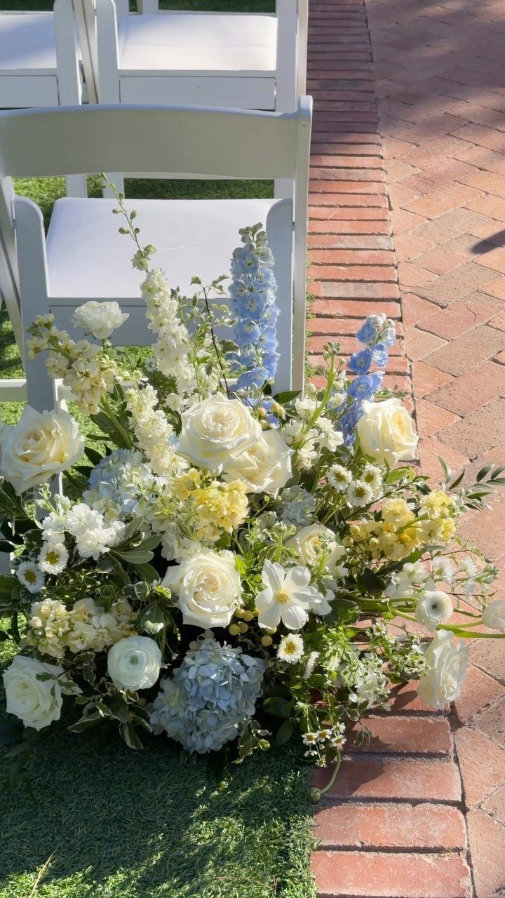 White, yellow, and light blue flowers arranged in a large bouquet next to white chairs.