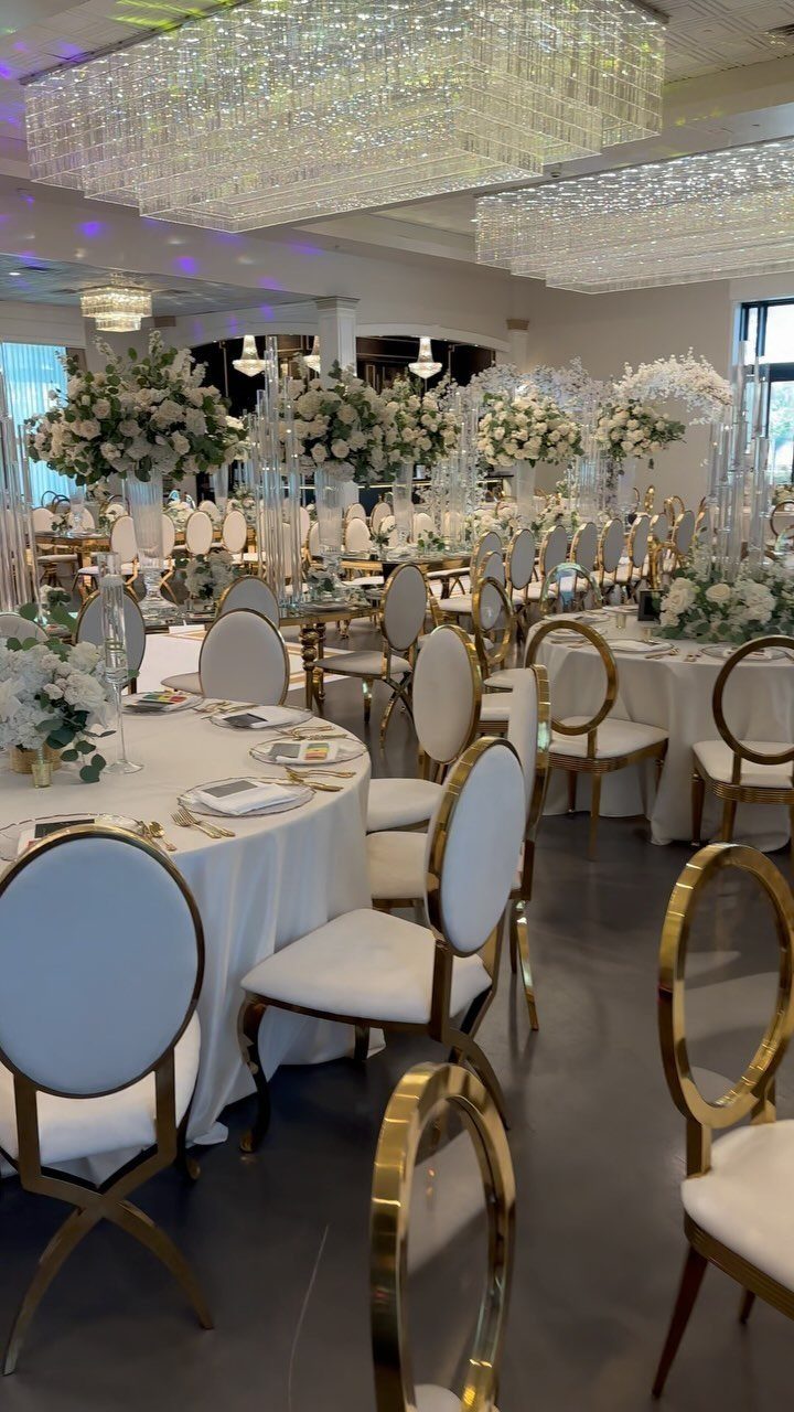 White wedding tables with gold chairs and large crystal chandeliers in a ballroom.