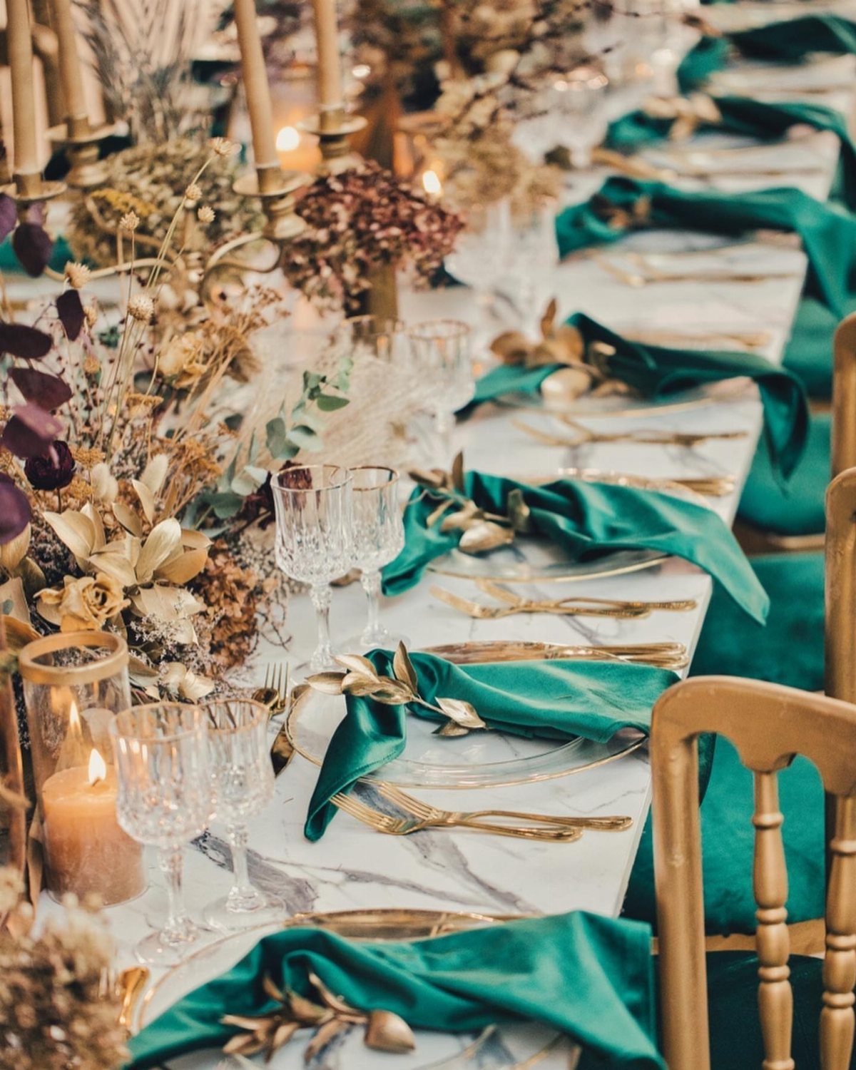 A wedding table setting featuring emerald green napkins, gold cutlery, and dried floral arrangements.