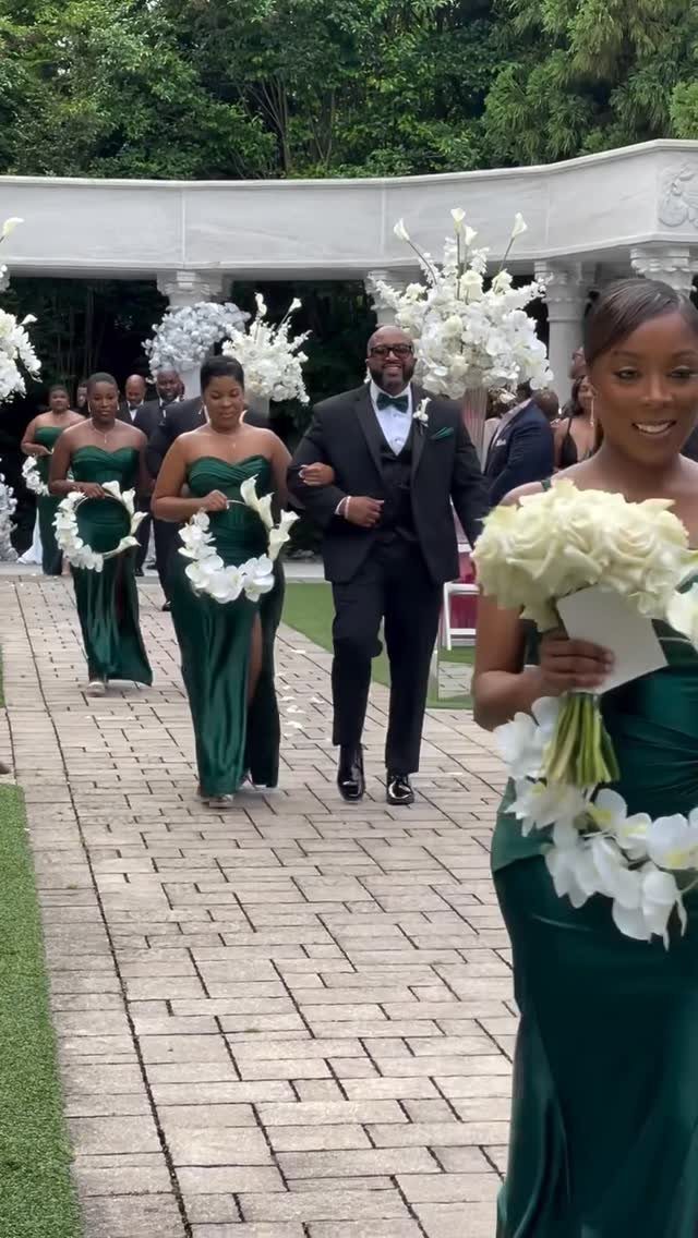 Bridesmaids in emerald green satin dresses walk down a stone path carrying white bouquets.