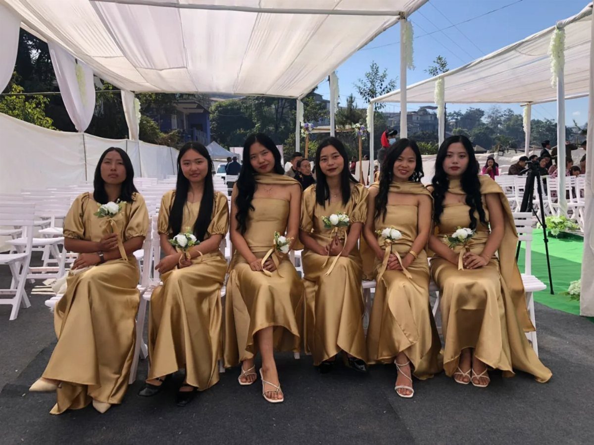 Six women in matching gold satin dresses sitting in a row holding white bouquets.