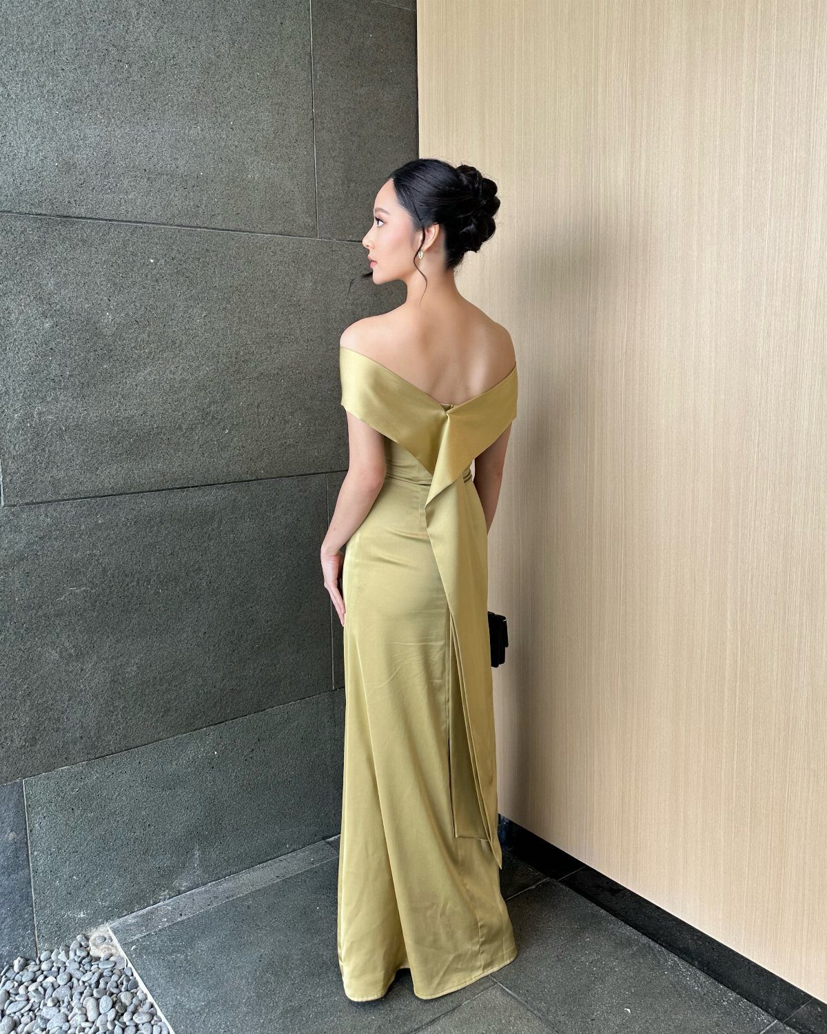 Woman in a long, off-the-shoulder olive green satin gown standing against a textured wall.