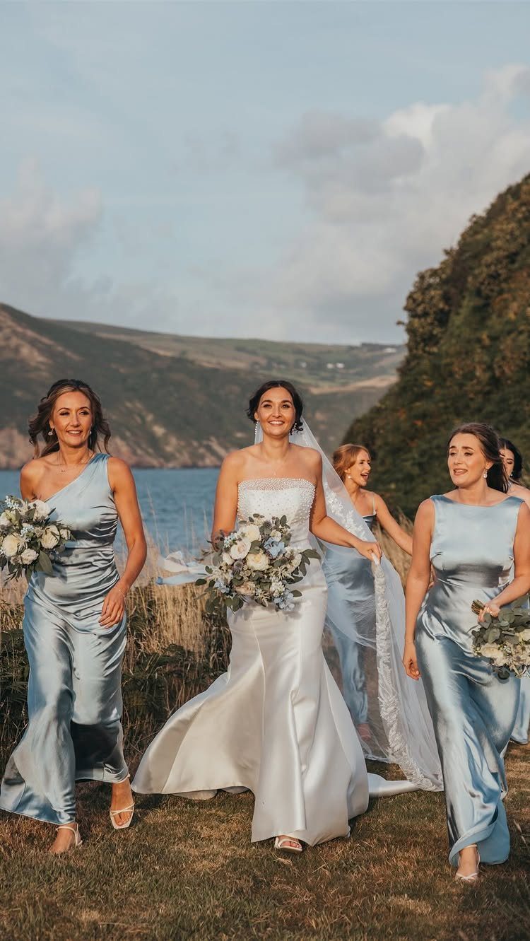 Bridesmaids in light blue satin dresses walking with a bride near the ocean.