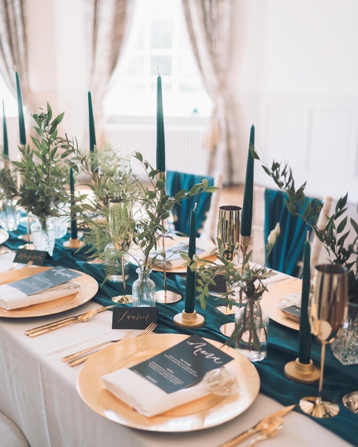 A wedding table setting featuring teal runners, gold plates, and tall teal taper candles.