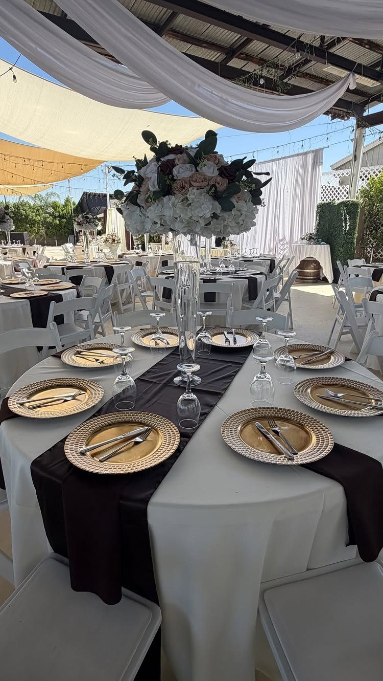 Wedding table setting featuring gold chargers, white linens, and a dark brown runner with floral centerpiece.
