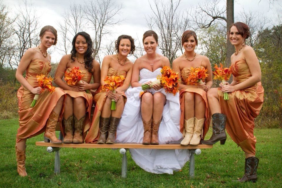 Bridesmaids in champagne and rust dresses and a bride sitting on a bench wearing cowboy boots.