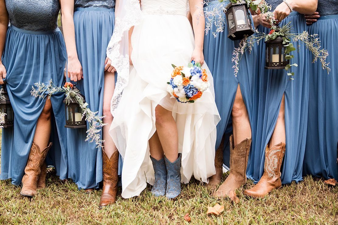 Bridesmaids in dusty blue dresses and cowboy boots standing next to a bride in white.