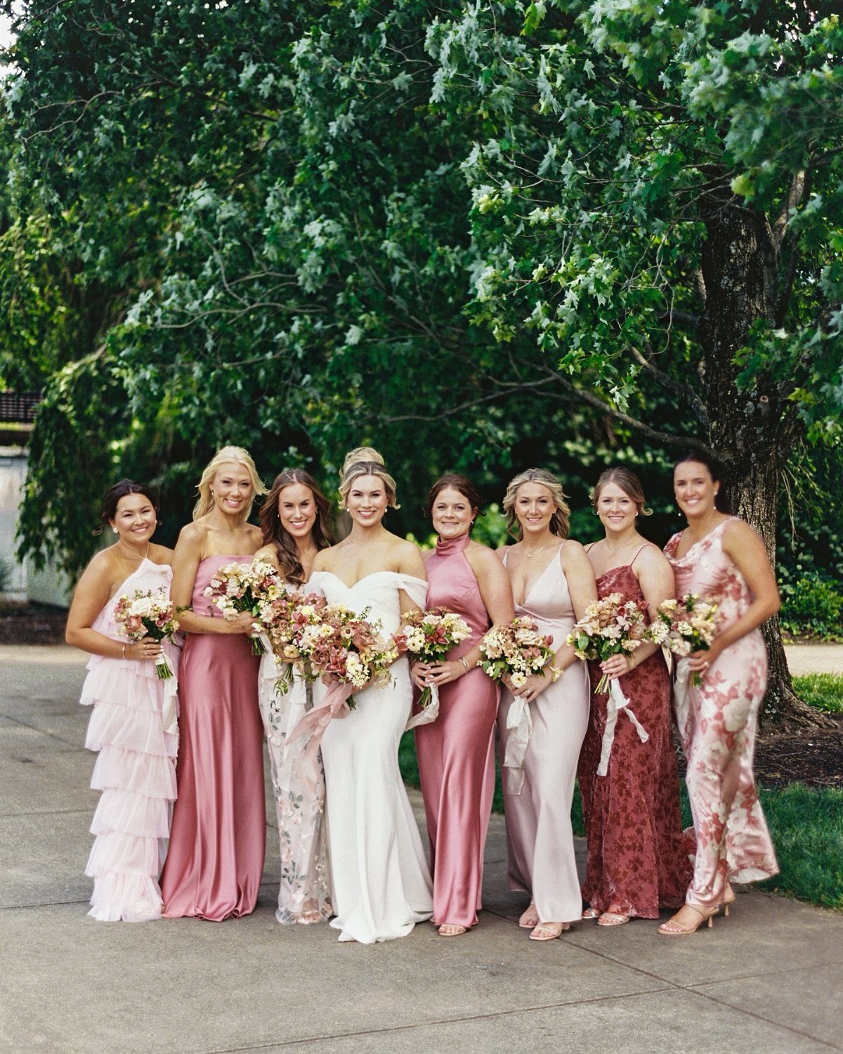 Nine women in various floor-length gowns standing together holding floral bouquets outdoors.