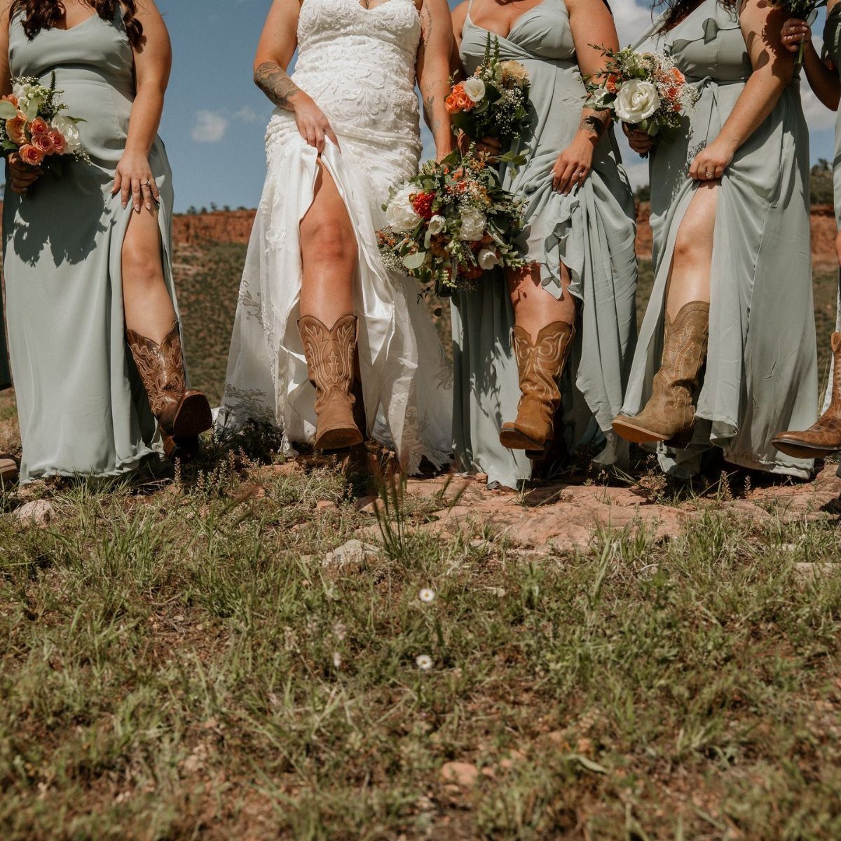 Bridesmaids in sage green dresses and a bride in white lace wearing cowboy boots outdoors.