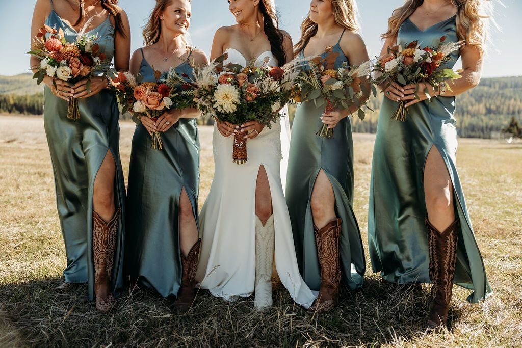 Bridesmaids in teal satin dresses with high slits and cowboy boots standing in a field.