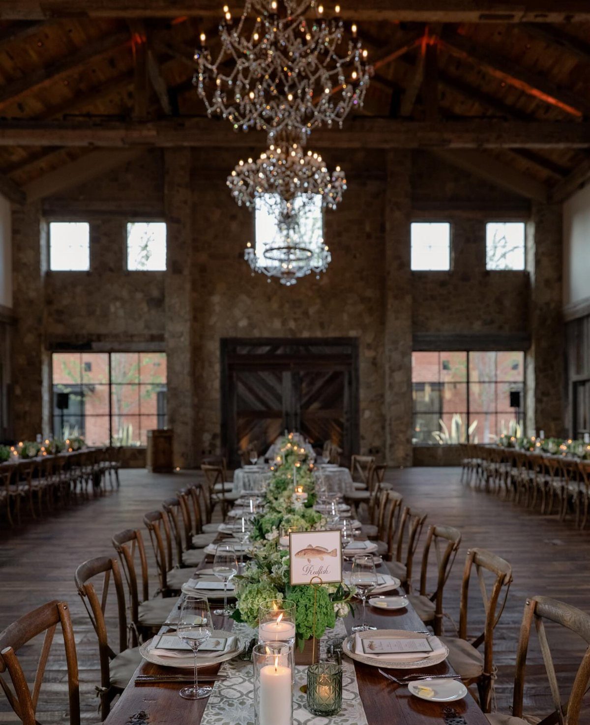 Long wooden wedding tables decorated with greenery and candles inside a rustic stone barn venue.