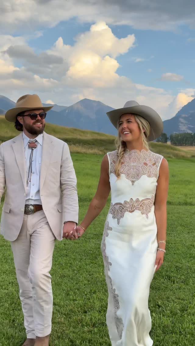 A couple in western-inspired wedding attire walking through a green field with mountains.