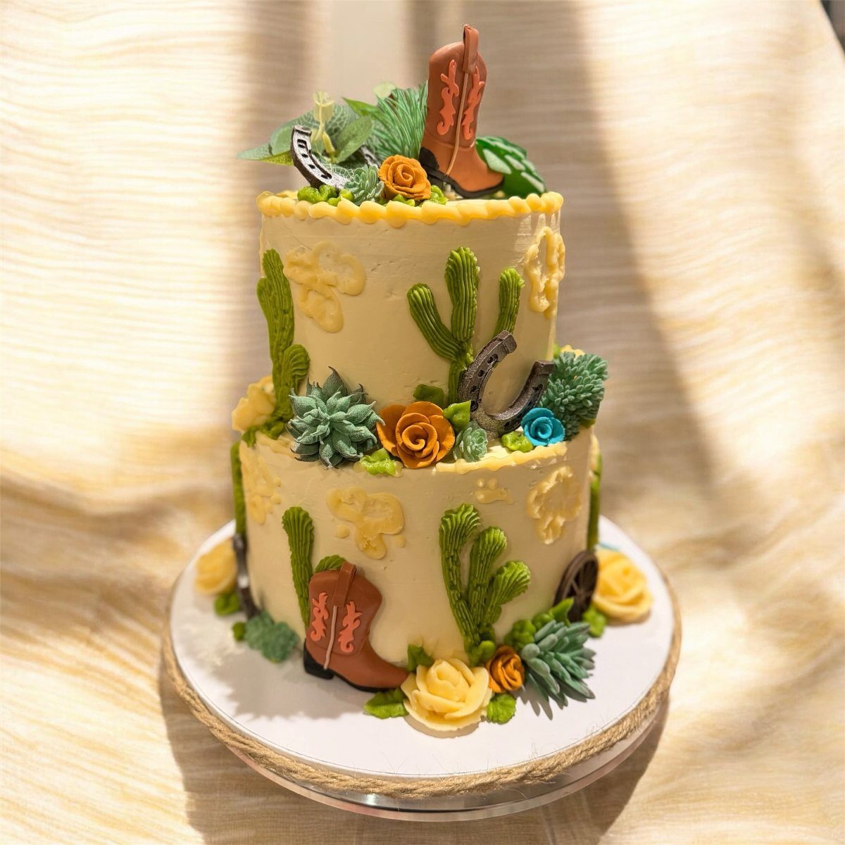 Two-tier cream cake decorated with cactus frosting, cowboy boots, roses, and horseshoe ornaments.