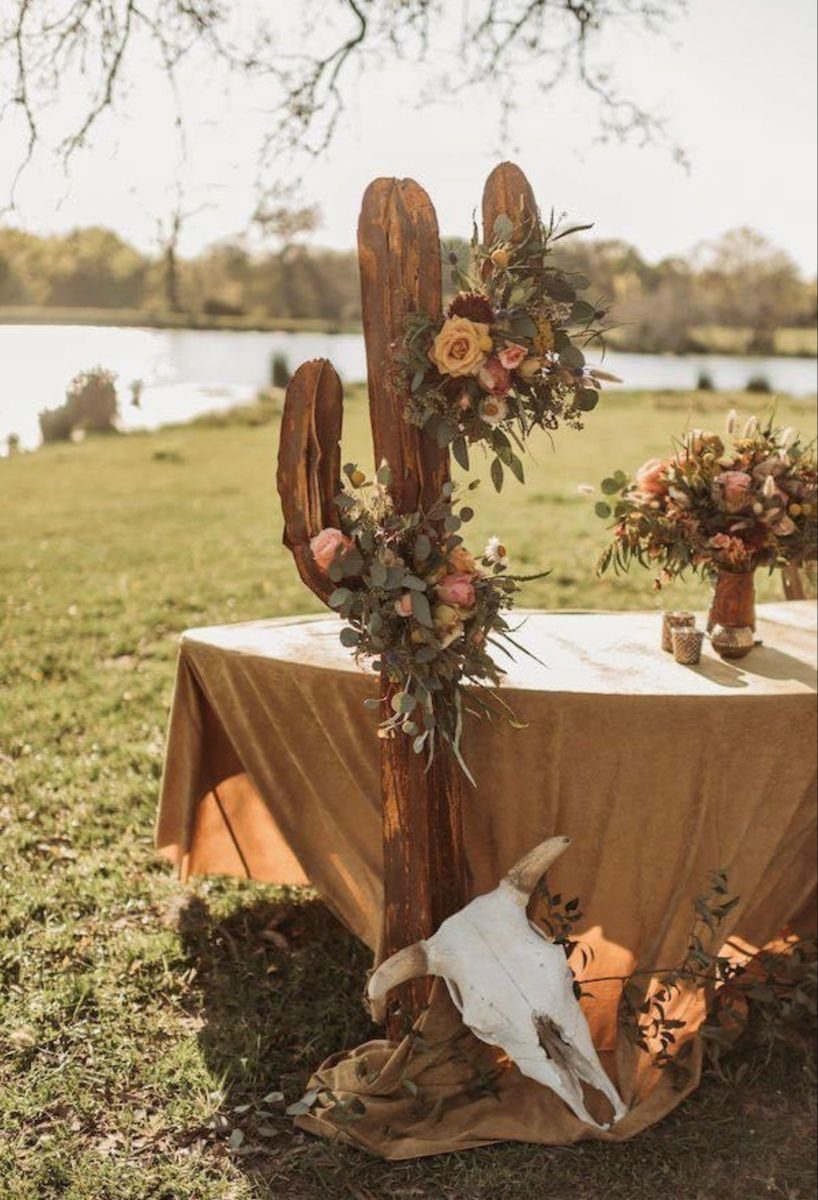 Wooden cactus sculpture with floral accents and an animal skull near a decorated table outdoors.