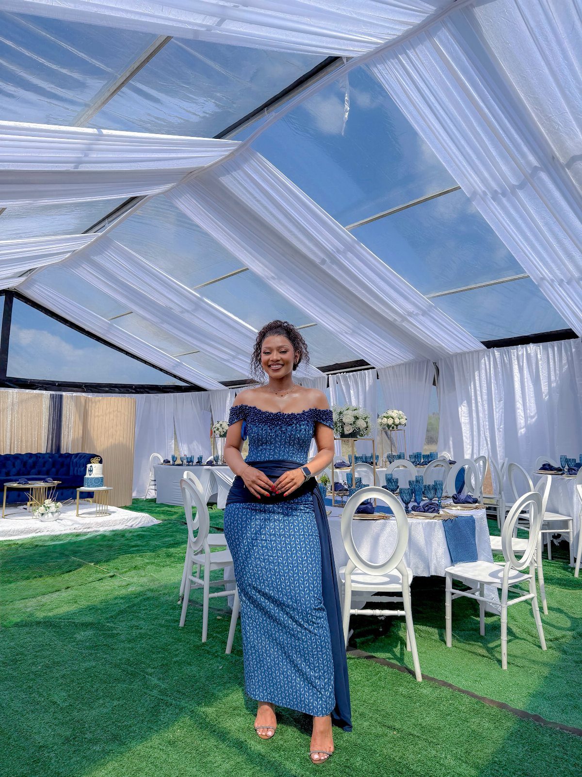 A woman wearing a blue patterned off-the-shoulder dress stands in a decorated outdoor event tent.