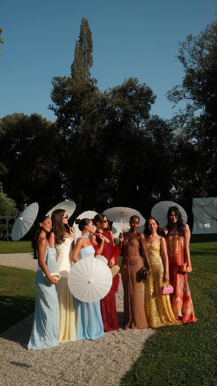 Seven women in colorful long dresses holding white parasols on a gravel path outdoors.