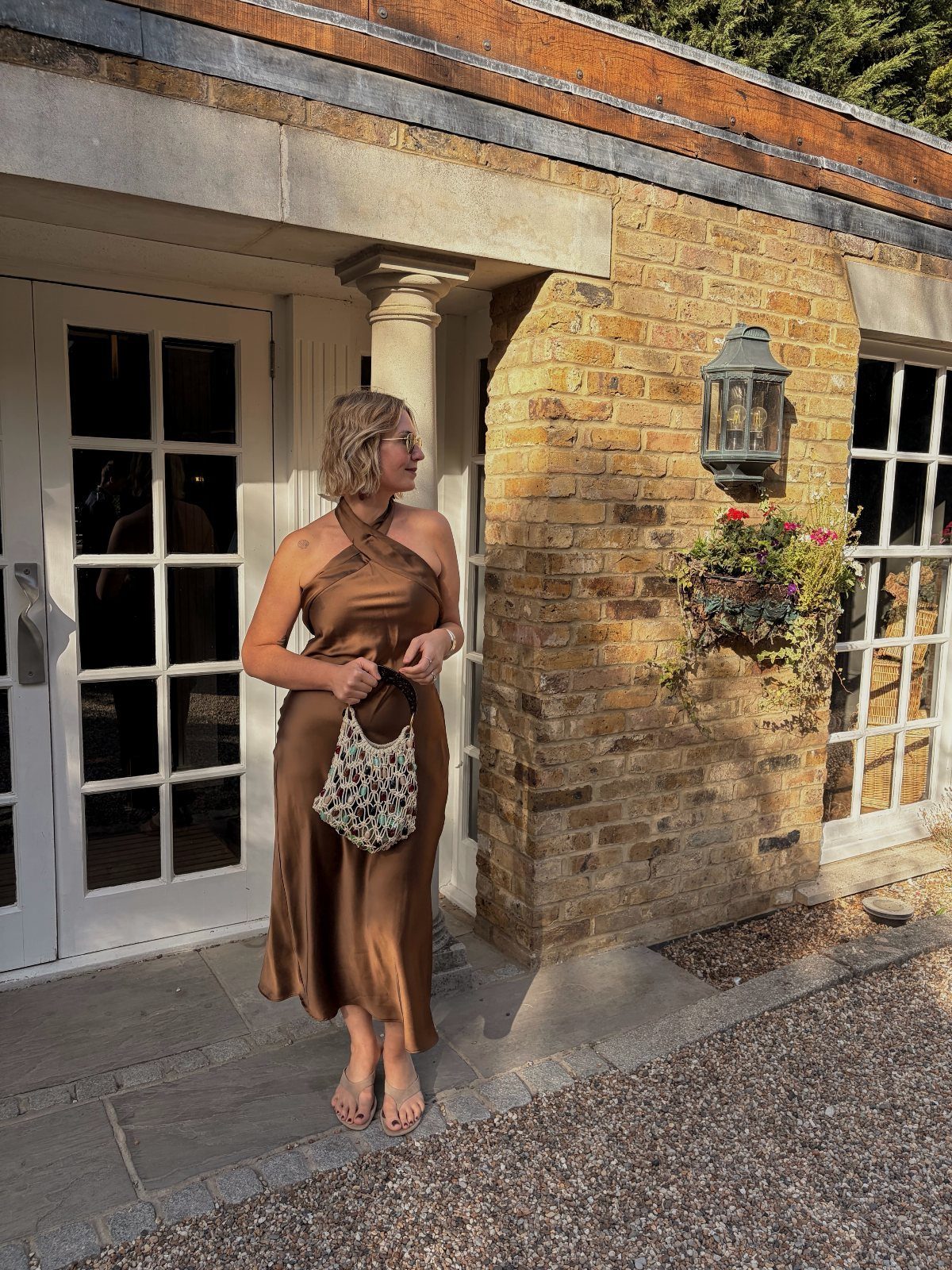 A woman wearing a bronze satin halter dress and holding a small patterned bag outdoors.