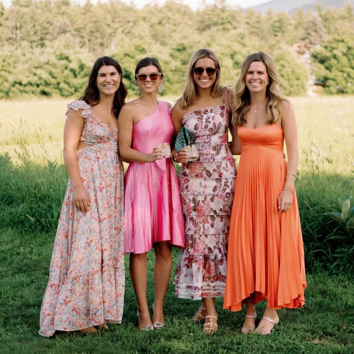 Four women in colorful summer dresses posing together in a grassy field with trees behind them.