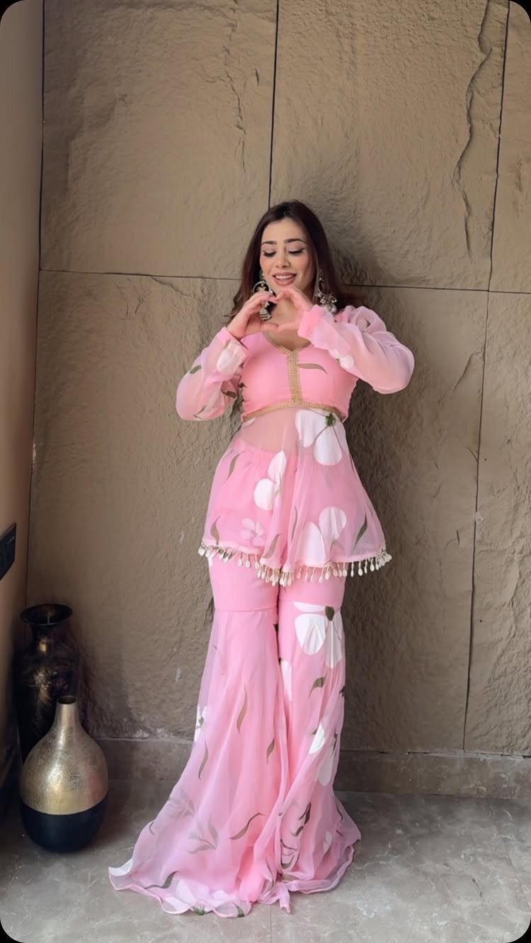 Woman posing in a pink floral two-piece outfit with wide-leg flared trousers against a stone wall.