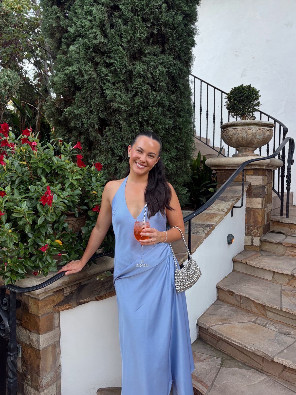 Smiling woman in a light blue halter maxi dress holding a cocktail on stone stairs.