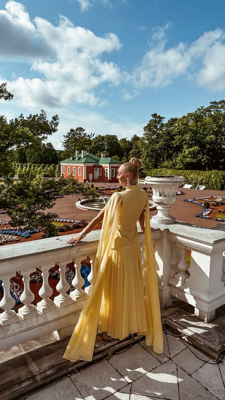 Woman in a flowing yellow formal gown standing on a white stone balcony overlooking a garden.