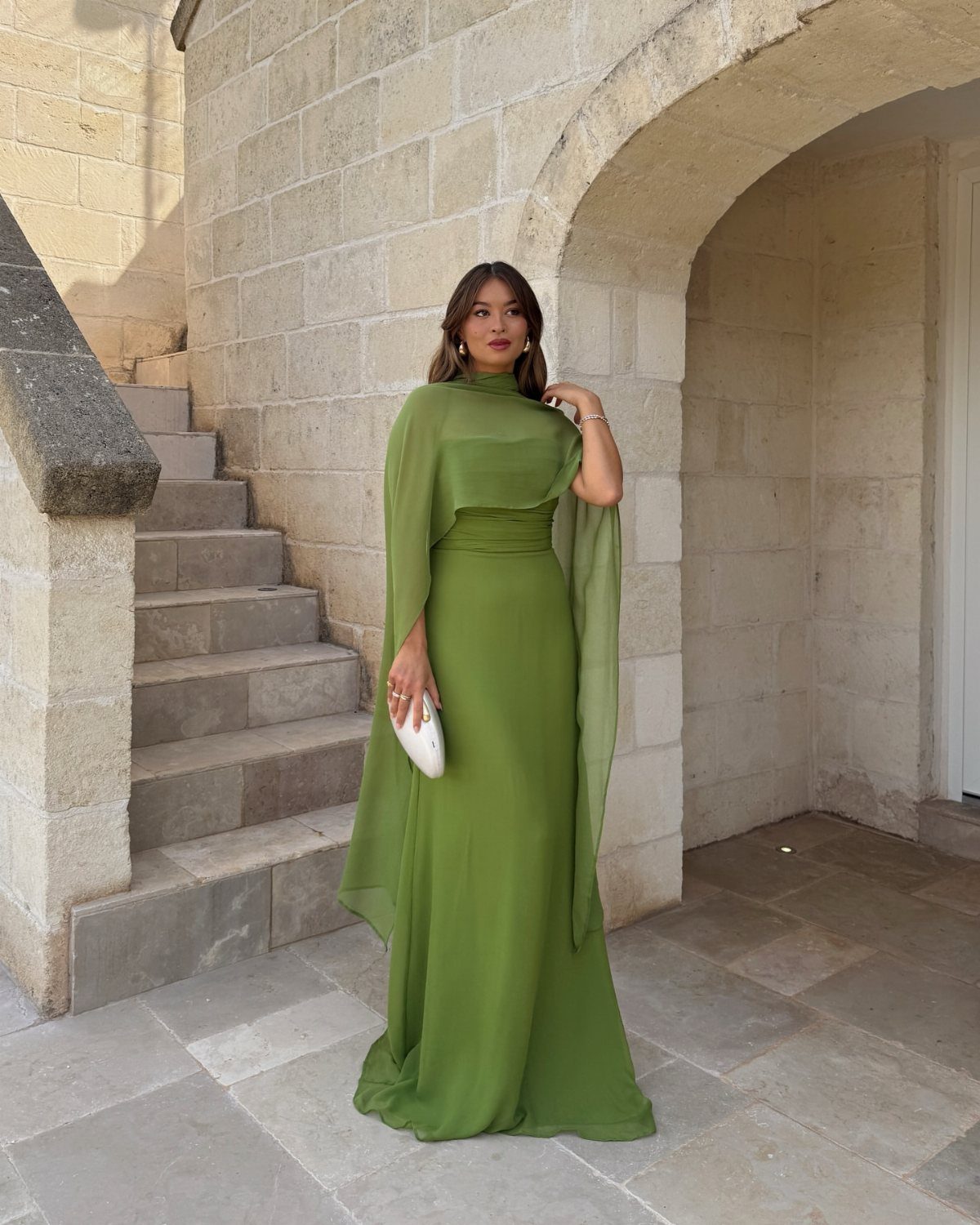 Woman in a long olive green chiffon gown posing by stone stairs and an archway.