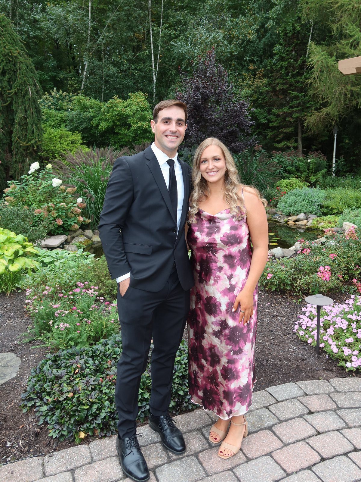 A man in a black suit and a woman in a patterned purple dress posing in a garden.