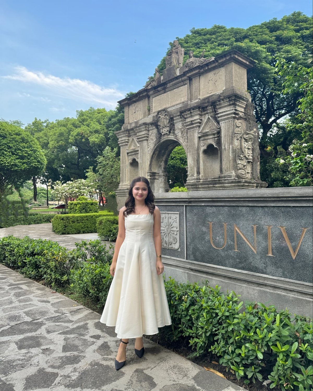 A woman in a white sleeveless A-line dress posing by a stone archway.